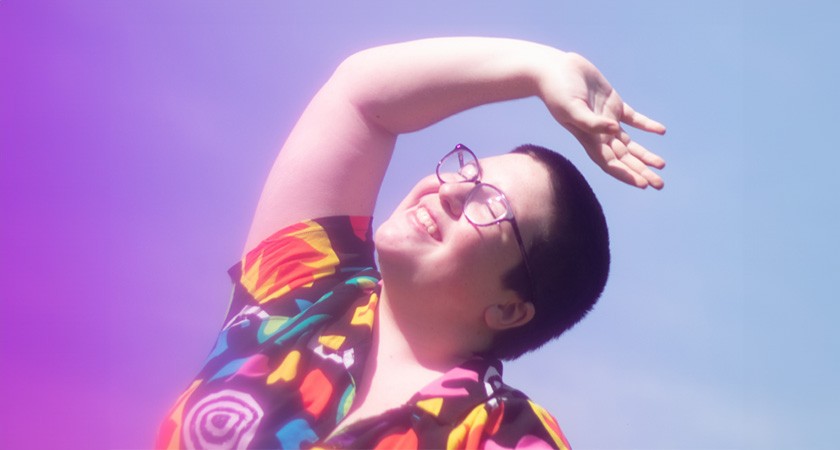 A white person with short dark hair, thin dark glasses, and a brightly colored pattern short-sleeved shirt smiles and squints as they raise their arm to block out the sun