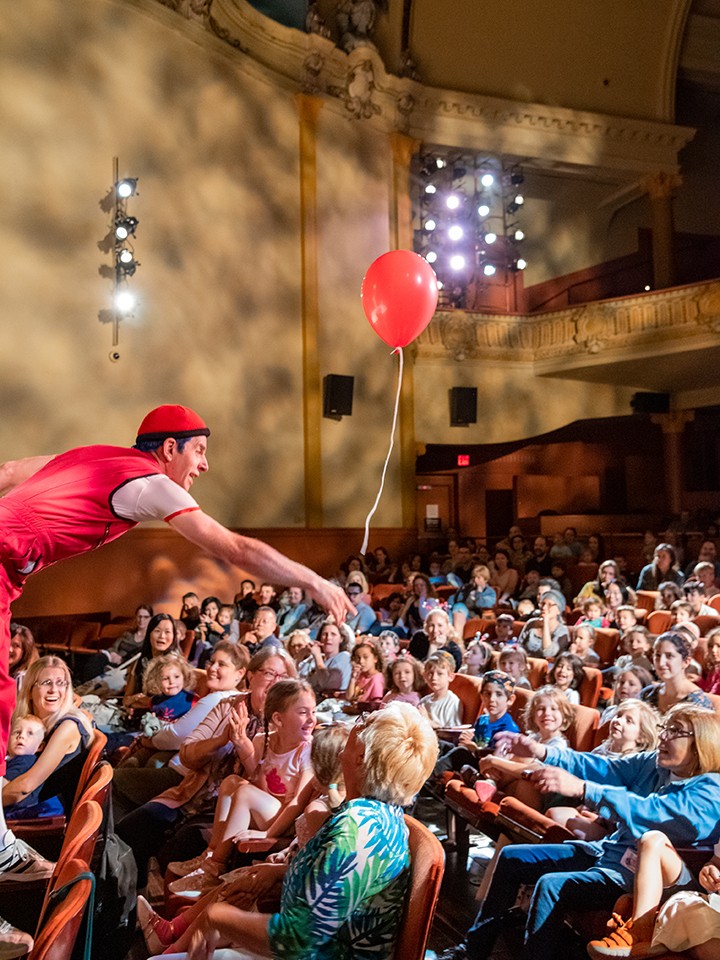 A person holds a baloon out to a group of families and children sitting inside a theater