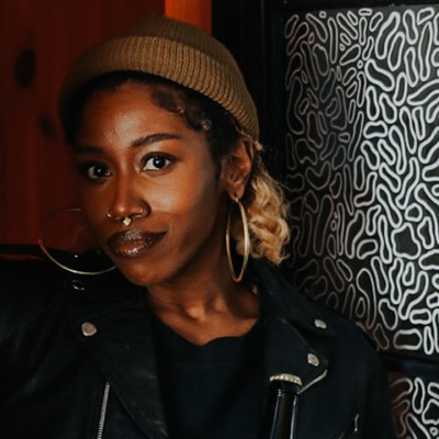 A woman with large hoop earrings, a black t-shirt, black leather jacket, and a light brown beanie smiles for the camera beside black-and-white abstract artwork