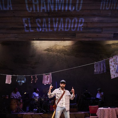 A man stands on stage. He's wearing a plaid shirt, light blue jeans, baseball cap, and has a guitar strapped around his back. A line of laundry is hung up behind him. He's holding his fingers upwards towards words projected including "El Salvador, Guatemala, Chanmico, Honduras"