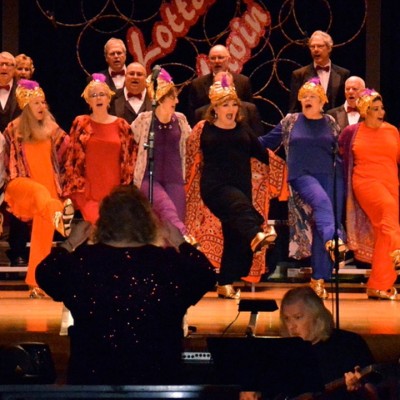 A group of older white dancers perform on a stage in front of a conductor and a person playing the guitar