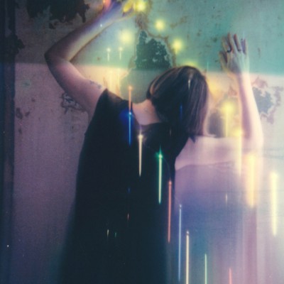 Artistic photograph that shows a white woman from behind with her face and arms pressed against a blue-and-pink tinted wall. She has short brown hair and is wearing a sleeveless black dress. The photo has been altered with streaks of colorful lights