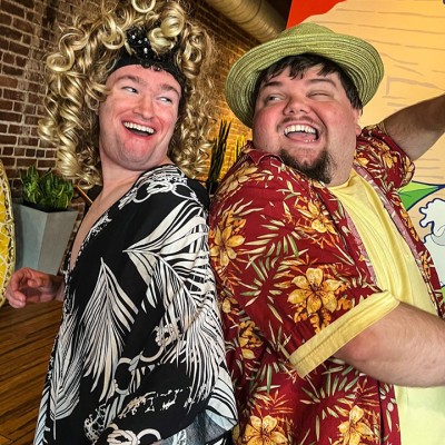 Two actors pose joyfully while wearing eccentric colorful tropical shirts. The actor on the left is wearing a curly blonde wig and the actor on the right is wearing a straw hat