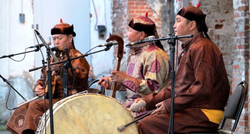 Three people wearing black headbands sit in seats in front of microphones while holding various instruments