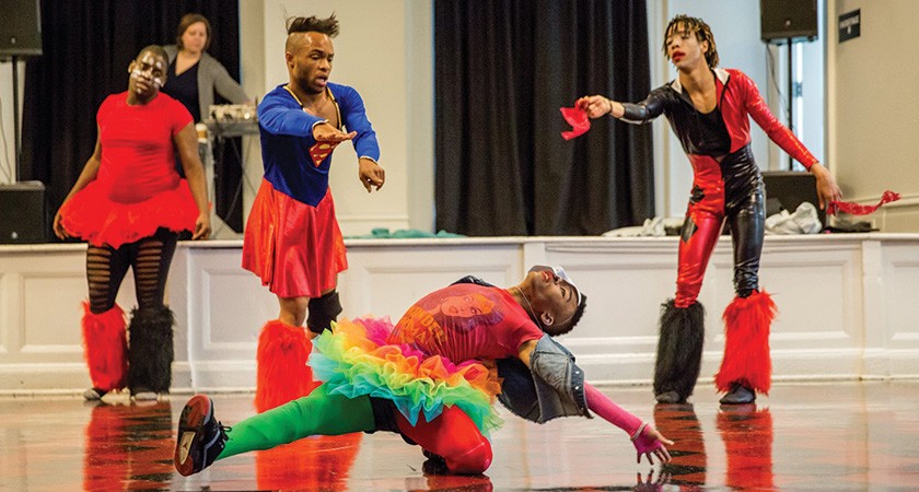 A racially diverse group of dancers in colorful theatrical dance costumes perform on a ballroom floor