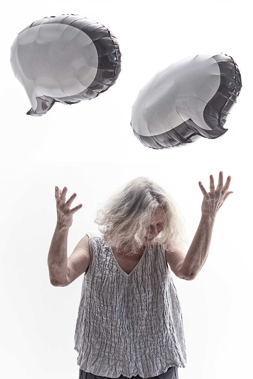 A white woman with a silver tank top laughs as she throws her arms in the air. Two silver word-shaped balloons hover above her head