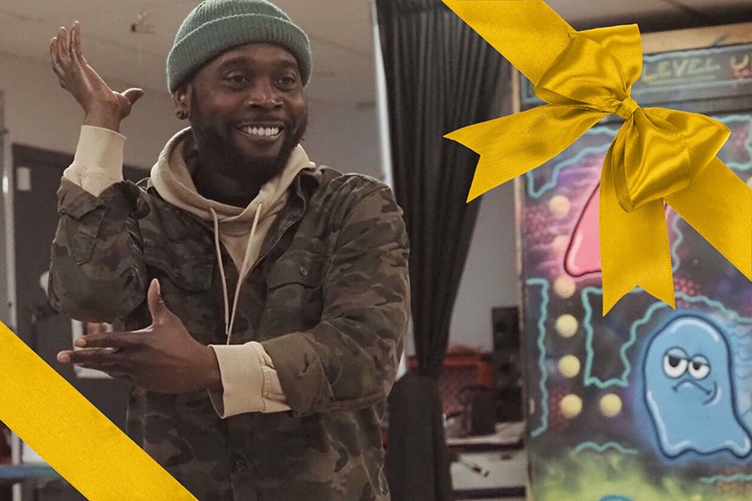 Photo of a smiling Black man wearing a beige hoodie, a camoflauged jacket, and a green beanie dancing inside an arts space. Photograph has been photoshopped onto a gift card frame