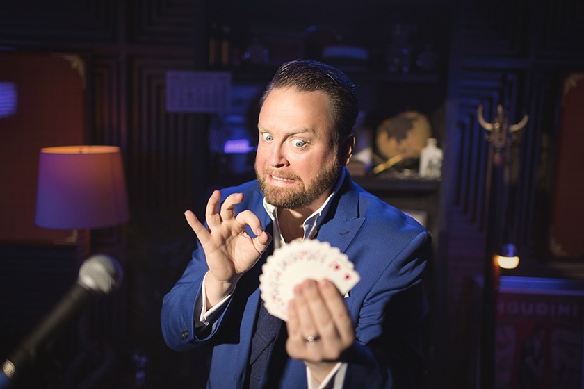 A white man with short dark hair and a brown beard and wearing a blue suit pretends to grimace nervously while reaching one of his hands out to pick out a card from a deck in his other hand