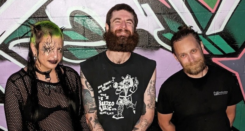 Three smiling people stand in front of a colorful graffiti wall. The person onthe left has green hair, black makeup, and is wearing a black bra under a blash mesh top. The person in the middle has a big brown beard and is wearing a sleeveless black punk top. The person on the right has a blonde beard and is wearing a black t-shirt