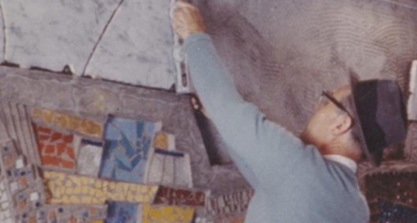 Faded photo of a white man with glasses, black and gray hat, and a blue sweater installing a colorful mural on a wall