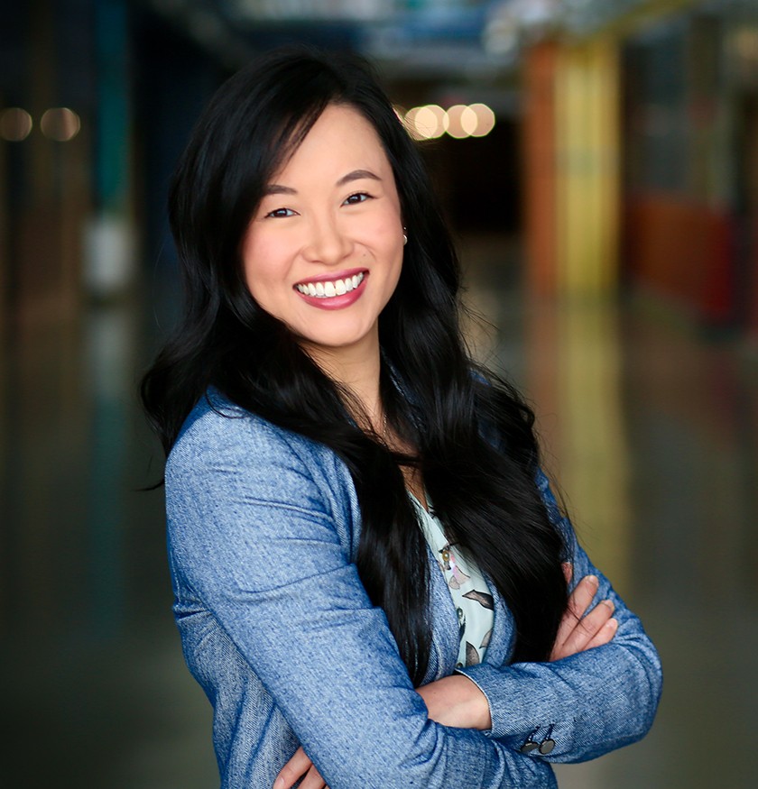 A smiling Asian woman with long straight black hair and a long-sleeved blue shirt stands with her arms crossed