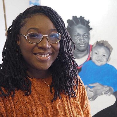 Smiling Black woman with long dark hair wearing gold glasses, nose ring, hoop earrings, and a bright orange sweater