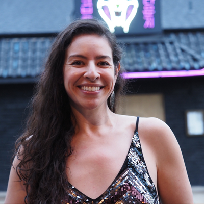 Mexican and American Woman with long curly brown hair, wearing a shiny black-and-silver tank top, smiles for the camera