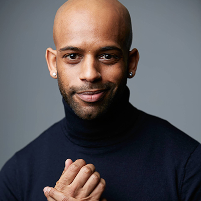 Smiling bald Black man with a closely-shaved dark beard, silver stud earrings, and a dark blue turtleneck