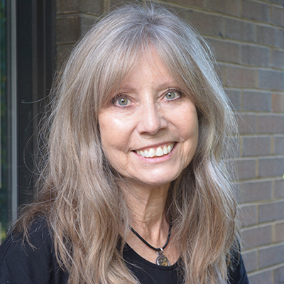 Smiling white woman with long straight grayish blonde hair, wearing a necklace with a black rope and silver pendant and a black shirt