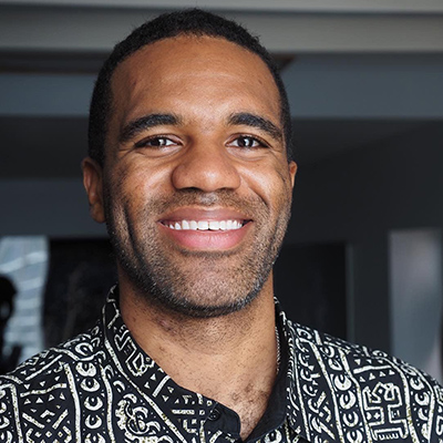 A smiling Black man with short dark hair, a closely shaved beard, and a black-and-white patterned shirt