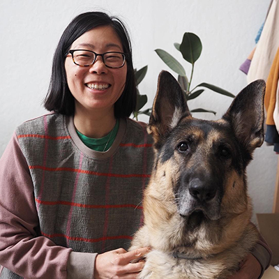 Smiling Asian woman with shoulder-length straight black hair wearing black glasses, and a green t-shirt under a purple, red, and gray long-sleeved shirt. She's posing next to a very big dog who's staring at the camera