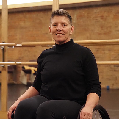 A white woman with short hair, a black long-sleeved shirt, and black pants smiles while sitting in a wheelchair