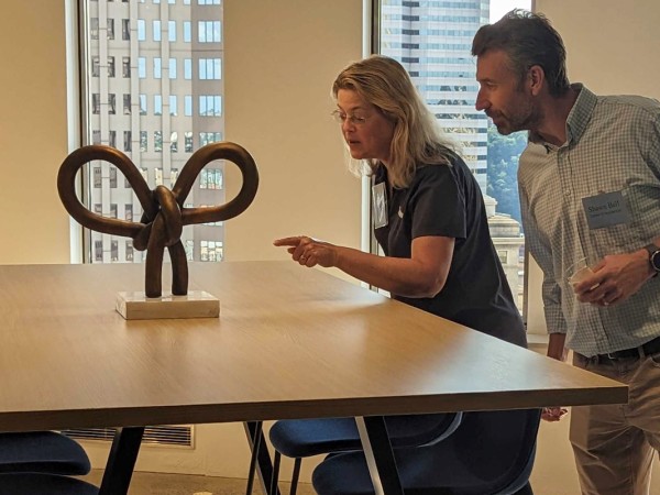 A couple leans over to look closer at an abstract metal sculpture sitting on a table in front of windows showcasing Downtown Pittsburgh hi-rise buildings