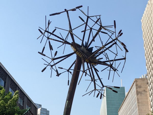 A metal sculpture of a dandelion shown photographed looking upwards. It's pictured in front of a bright blue sky, with high-rise Downtown buildings pictured in the background
