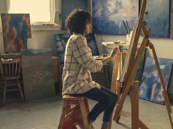 A person with curly shoulder-length black hair, wearing a plaid shirt and jeans, paints at an easel