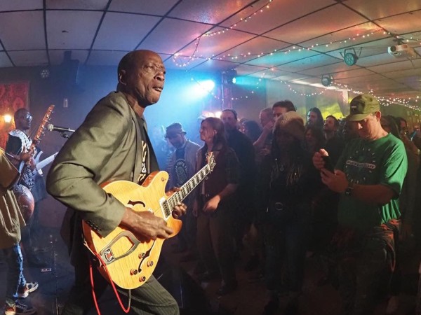 A bald Black man wearing a suit jacket, black t-shirt, and striped slacks plays an electric guitar on stage in the foreground. Two musicians are pictured behind him on stage, while a large crowd is pictured to the right of the photograph