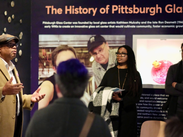 A man in a tan coat and hat speaks to a small group indoors; two people stand nearby listening, with photos and text displayed on the wall behind them. Some backs of audience members are visible in the foreground.