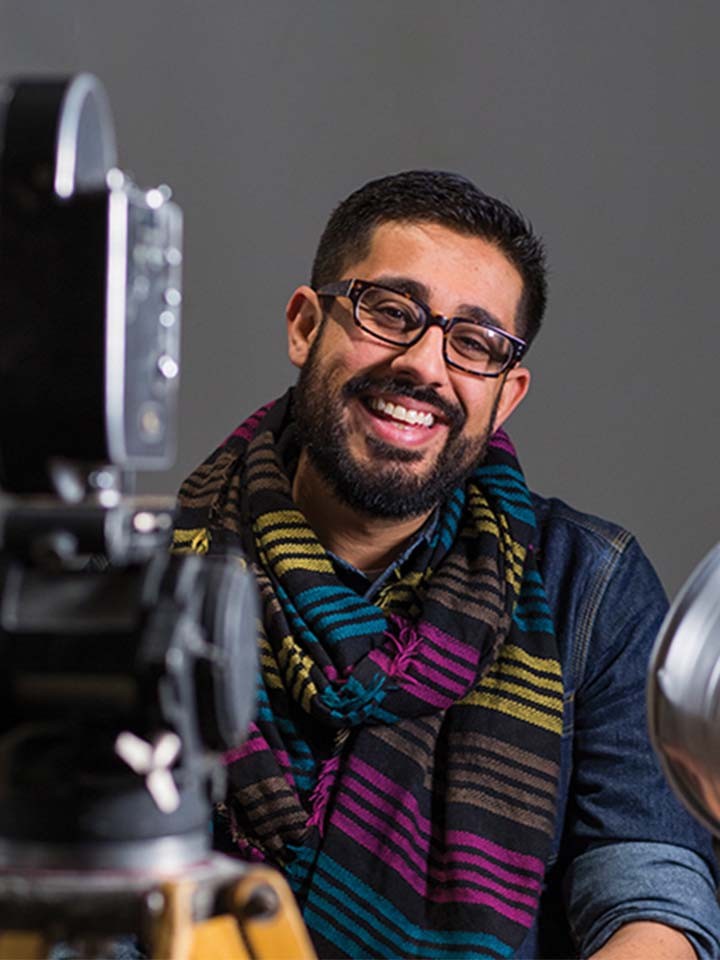 An image of Adil Mansoor, a theatre director and educator centering the stories of queer folks and people of color. He has brown skin and dark hair, a beard, and black glasses. He sits in front of a movie camera and light, wearing a striped, colorful scarf.