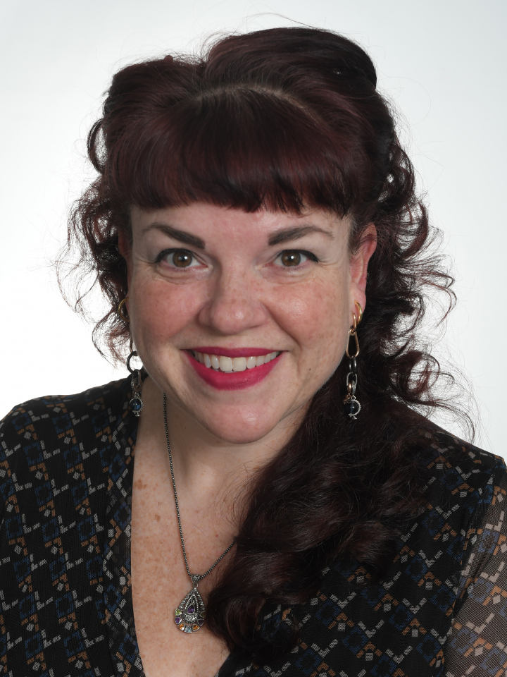 Smiling woman with dark auburn hair styled in a high curled updo, wearing red lipstick, dangling earrings, and a dark patterned top