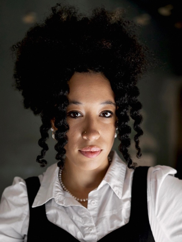 Portrait of a person with curly dark hair, wearing a white button-up shirt and dark vest