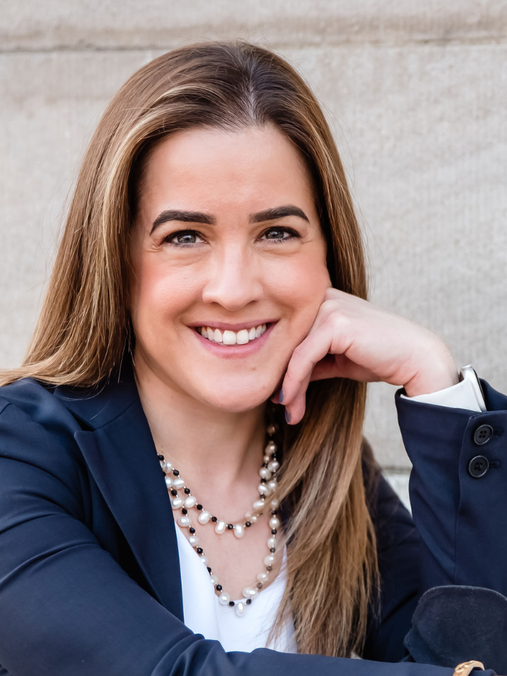 Smiling woman with long straight hair wearing a navy blazer and layered pearl necklace