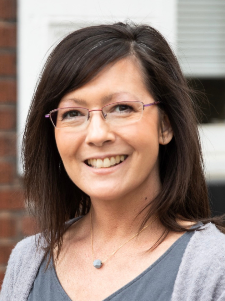 Smiling person with dark shoulder-length hair and rectangular purple-rimmed glasses, wearing a gray cardigan and a small heart-shaped pendant necklace