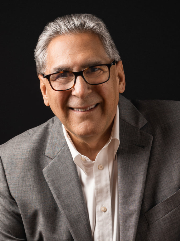 Smiling man with short gray hair wearing black rectangular glasses and a gray blazer over a white button-up shirt