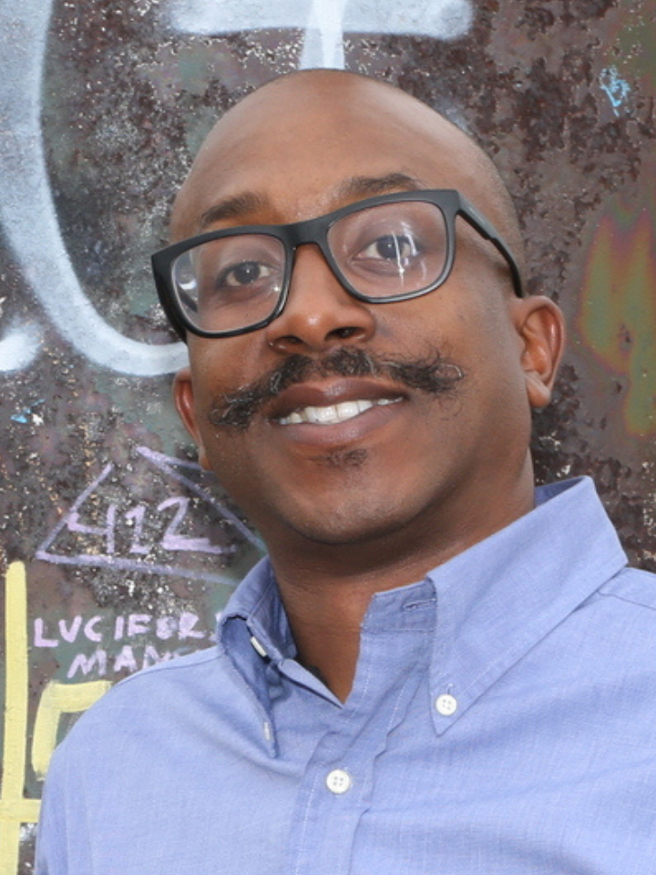 Smiling man with a dark mustache, black glasses, wearing a blue button-down shirt