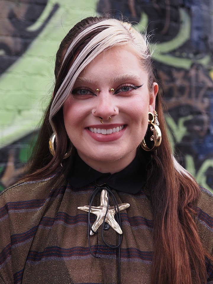 Smiling white person with long dark hair with chunky white streaks, multiple nose and ear piercings, wearing a brown striped shirt and a large metallic star necklace