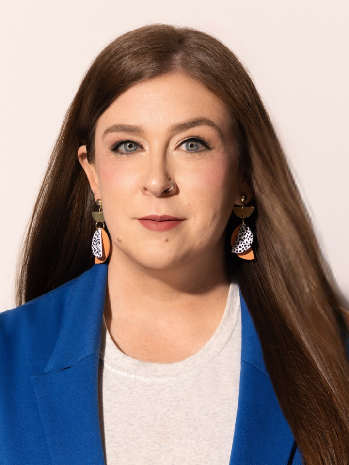 ortrait of a person with long brown hair, wearing blue outerwear over a white top, and dangling earrings