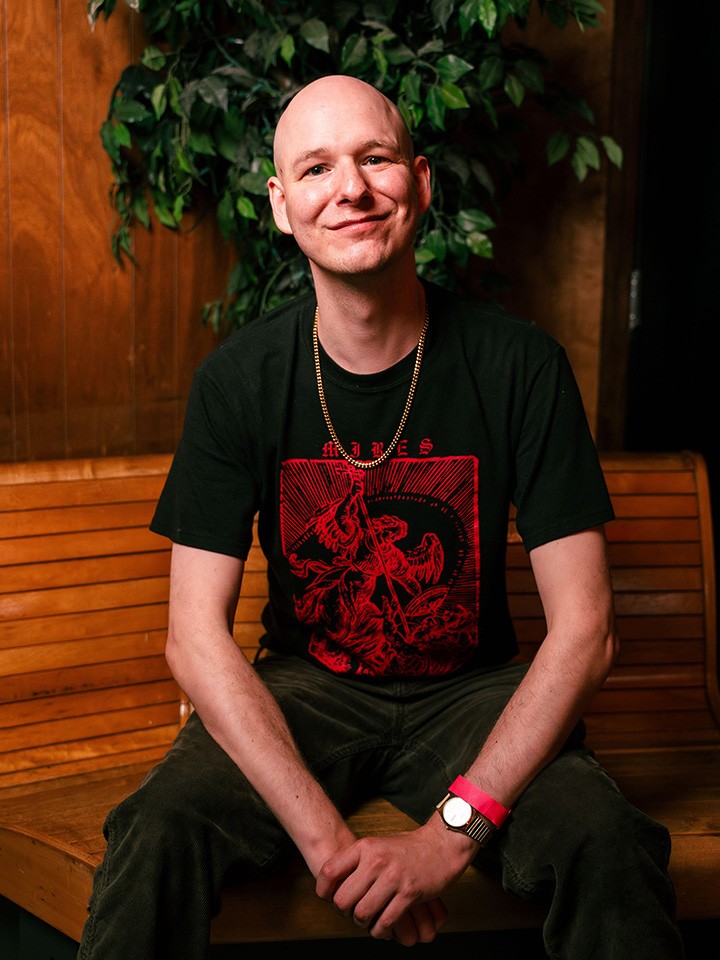A smiling person wearing a black t-shirt with a red graphic, a gold chain, and a wristwatch sits on a wooden bench in front of a wooden wall and leafy green plant.