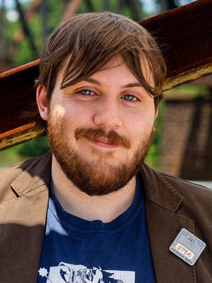 Close-up of a person with short brown hair and a full beard, wearing a dark jacket with a small name tag and a blue T-shirt