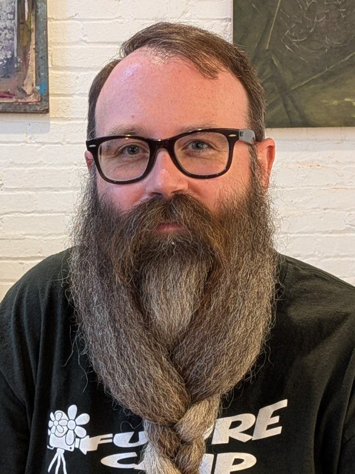 portrait of a person with dark-framed glasses and a long full beard, wearing a black T-shirt with white graphic text