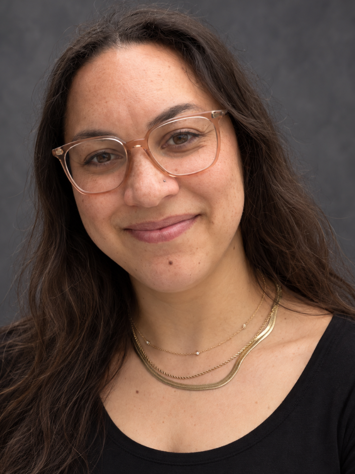 Close-up studio portrait of a person with long dark hair, wearing clear-framed glasses, a layered gold necklace, and a black top