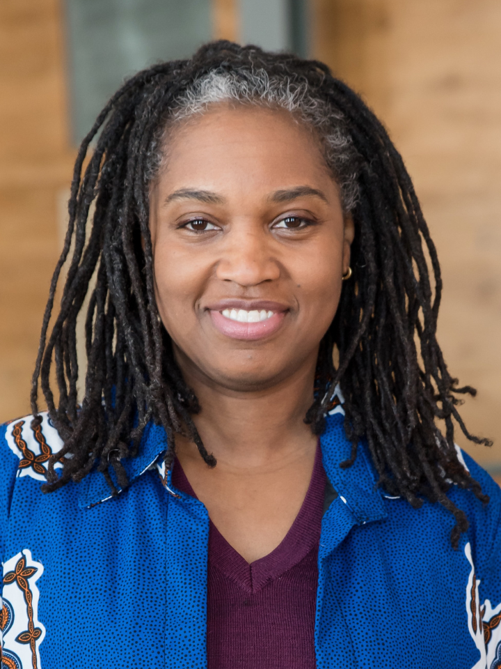A smiling person with shoulder-length black locs wearing a bright blue jacket over a dark maroon top
