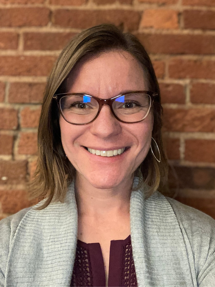 Smiling person with dark-framed glasses and shoulder-length hair, wearing a light gray cardigan over a dark maroon top