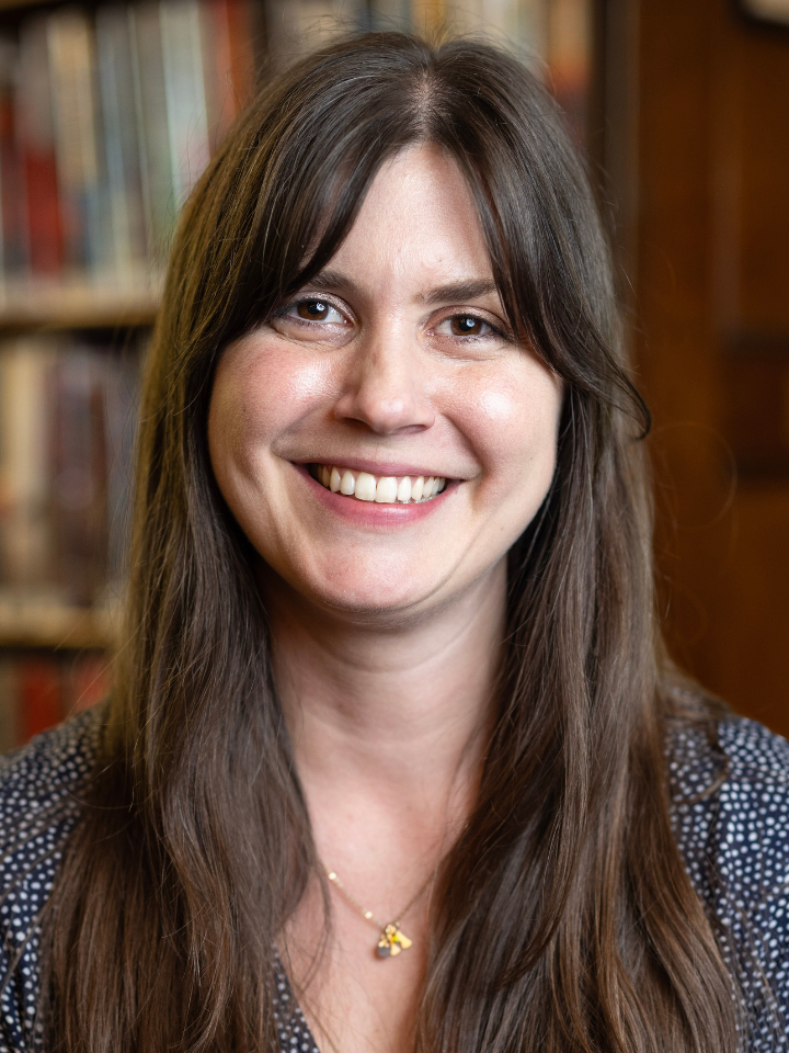 Smiling person with long dark brown hair, wearing a dark patterned top and a small gold necklace
