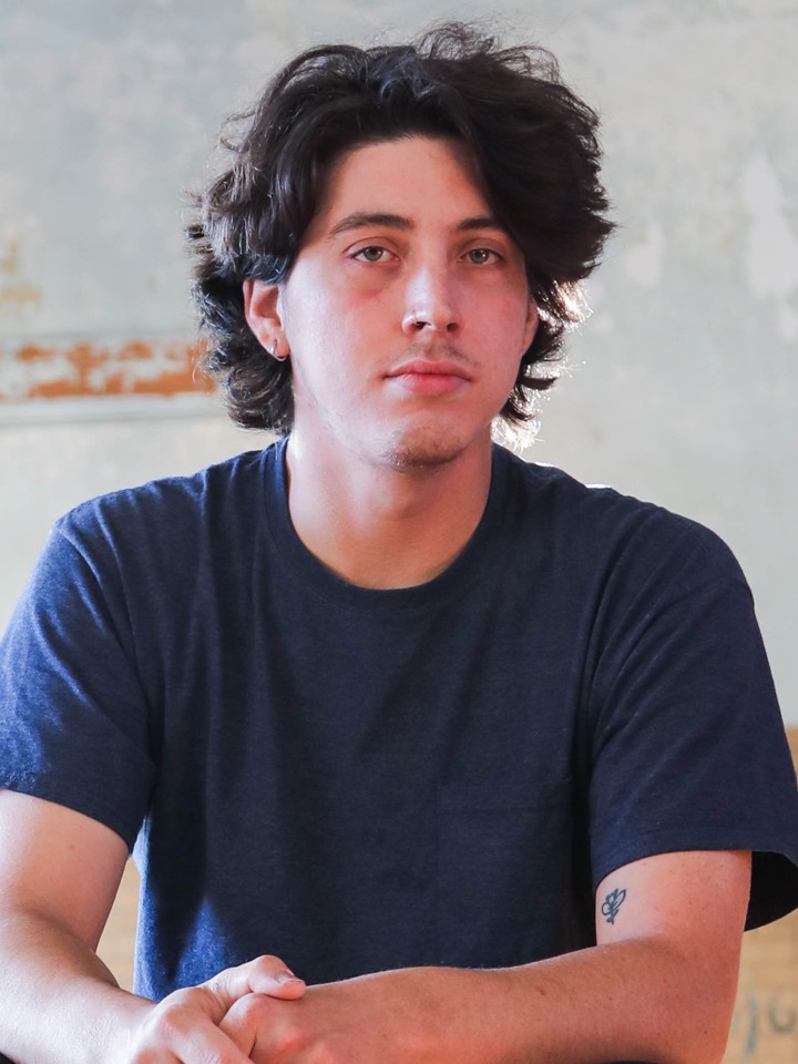 Latino person with short wavy dark hair, blue t-shirt, and a small tattoo on their arm
