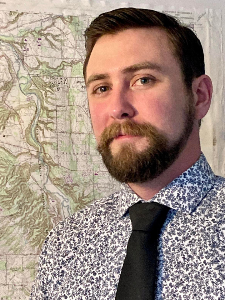 Portrait of a man with short dark hair and a full beard wearing a blue patterned shirt and black tie