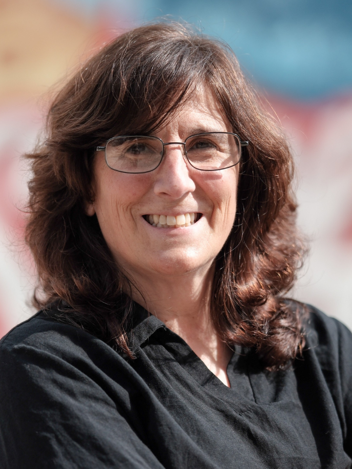 Smiling person with shoulder-length dark brown curly hair and rectangular glasses, wearing a dark collared top