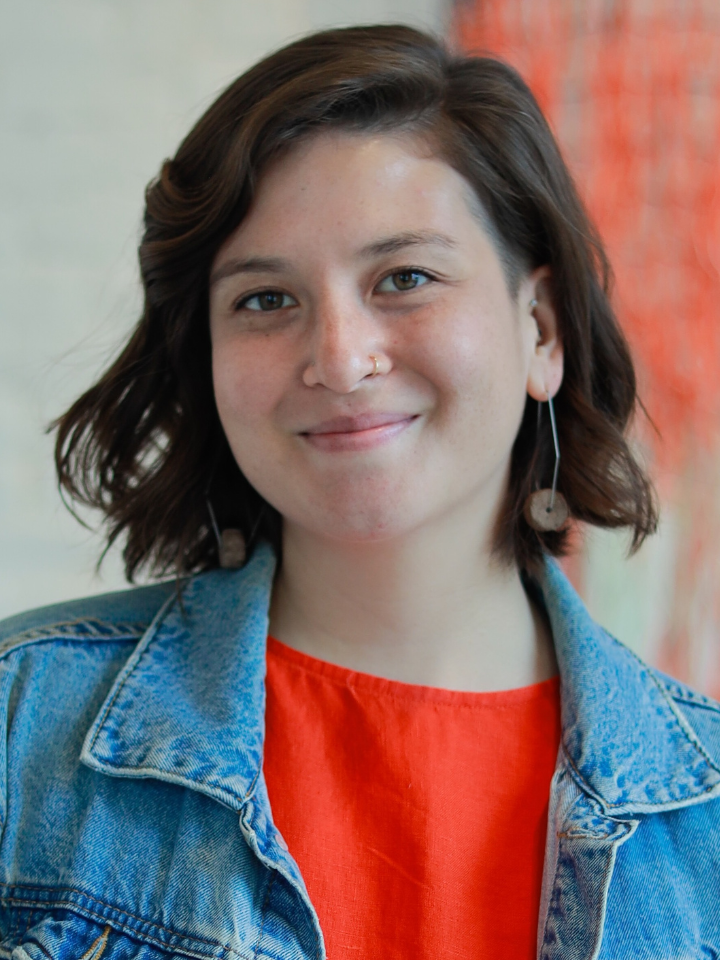 Portrait of a smiling person with shoulder-length dark hair wearing a blue denim jacket and an orange top