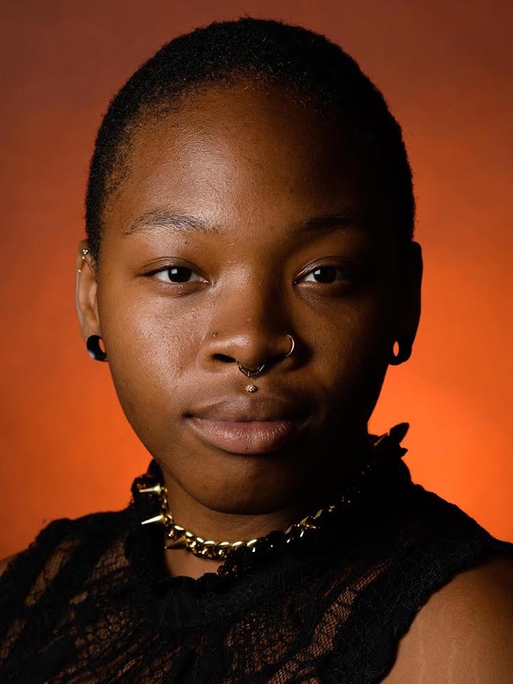 A Black person smiles for the camera. They have short black hair, black earrings, a silver chain choker, and a sleeveless black lace top. They're posed in front of a bright orange background