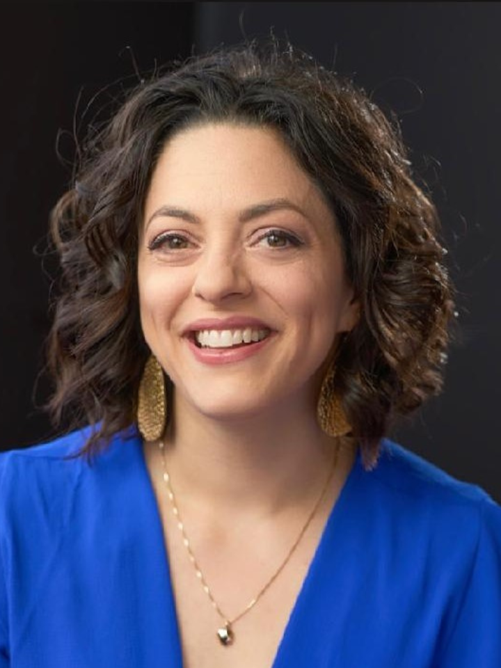 Smiling person with shoulder-length wavy dark hair wearing a bright blue top, gold hoop earrings, and a thin gold necklace