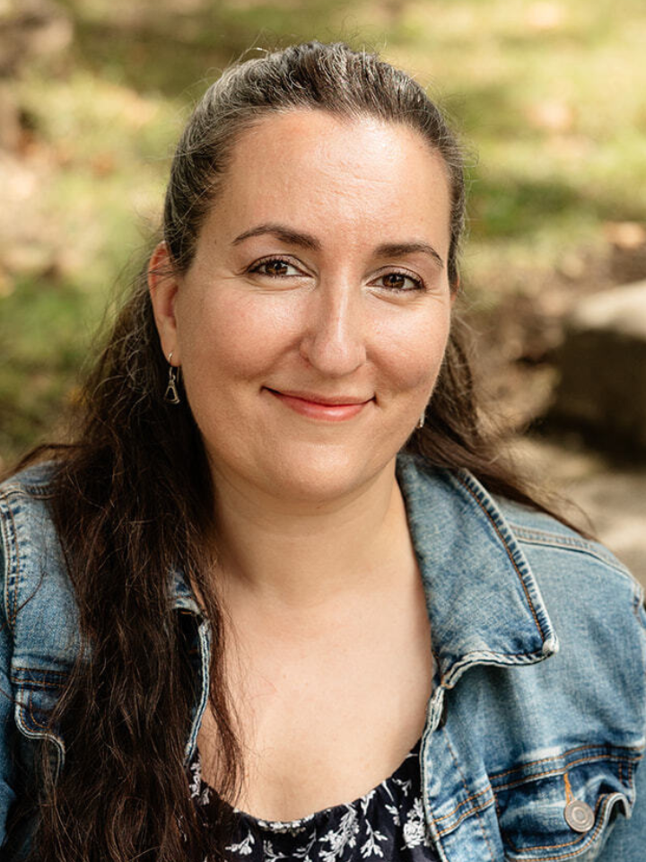 Smiling person with dark hair pulled back, wearing a denim jacket over a dark top